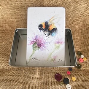 以下が含まれることがあります： 白い蓋が付いた銀色の金属製の缶。蓋には、紫色の花に止まっているミツバチの水彩画が描かれている。缶は開いており、蓋は缶の縁に置かれている。