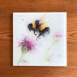 May include: Square glass art piece with a watercolour painting of a bumblebee and purple thistle flowers. The bee is black and yellow. The artwork is signed in the bottom left corner. The piece is on a wooden surface.