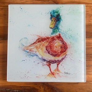 May include: Square glass coaster with a watercolour painting of a duck. The duck has a green head, a yellow beak, and a body with red, orange, and yellow hues. The artwork has a splattered paint effect, with the duck standing on a white background.