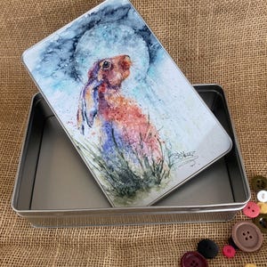 Puede incluir: Una lata de metal con una ilustración de acuarela de una liebre marrón mirando hacia arriba a una luna llena. La lata está abierta y la tapa está encima de la lata.