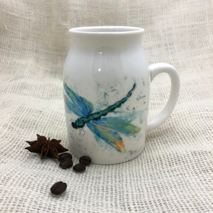 May include: White ceramic mug featuring a blue and green dragonfly design. The dragonfly is painted in a watercolour style with splashes of yellow and orange.