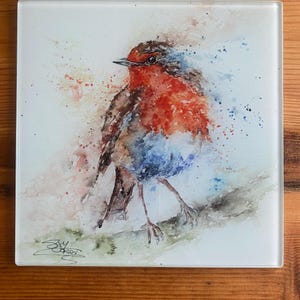 May include: Square glass coaster with a watercolour painting of a robin. The bird has a red breast, brown back, and blue accents. The artwork is signed in the lower left corner. The coaster is set against a light background.