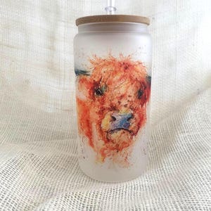 Puede incluir: Un vaso de vidrio esmerilado con tapa de bambú y pajita transparente. El vaso presenta una pintura de acuarela de la cara de una vaca de las Highlands, con tonos naranja, rojo y azul. El vaso está diseñado para bebidas frías.
