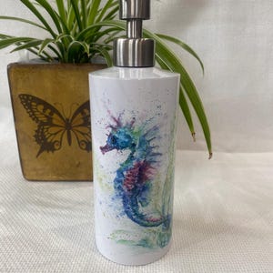 May include: White cylindrical soap dispenser with a stainless steel pump. The dispenser features a watercolour seahorse design in shades of blue, purple, and green. The seahorse is surrounded by colourful paint splatters.
