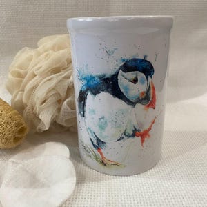 May include: White ceramic tumbler featuring a watercolor painting of a puffin bird. The bird has a black head, white body, and a vibrant orange beak. Blue and teal watercolor splashes surround the bird. The tumbler is cylindrical and sits on a white surface.