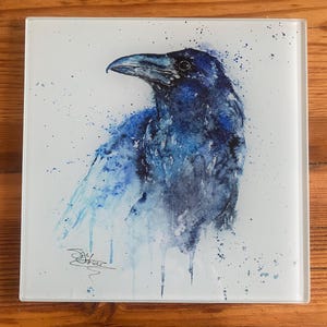 May include: Square glass art piece featuring a watercolor painting of a raven. The bird is depicted in shades of blue, with a detailed beak and feathers. The background has blue paint splatters and a signature.