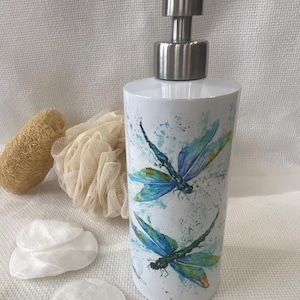 May include: White ceramic soap dispenser with a stainless steel pump, decorated with blue and green dragonfly illustrations. The dispenser is cylindrical and sits next to a loofah, a mesh sponge, and cotton pads.