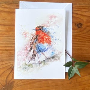May include: Watercolor painting of a robin bird on a white card with an envelope. The bird has a red breast, brown wings, and blue accents. The artwork is on a white card with a white envelope. A sprig of green leaves is on the right.