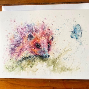 Puede incluir: Una pintura de acuarela de un erizo rosa con una mariposa azul volando cerca. El erizo está rodeado de salpicaduras de acuarela verde y marrón.