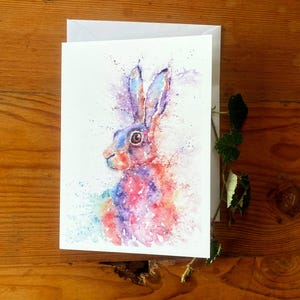 May include: Watercolor painting of a hare with blue, purple, red, and orange hues. The artwork is on a white card with an envelope, set against a wooden background. The hare's fur is depicted with colorful splashes.