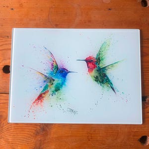 May include: A glass cutting board with a watercolor painting of two hummingbirds in flight. The hummingbirds are painted in shades of green, blue, red, and purple. The background is white.