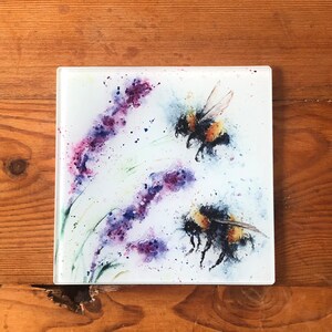 May include: Square glass coaster with a watercolour design of two bumblebees and lavender. The bees are black and yellow, with transparent wings. The lavender is purple and pink. The background is white, with splashes of colour.