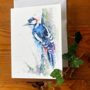 May include: A watercolor greeting card featuring a blue, black, and white woodpecker perched on a tree trunk. The bird has a red head marking and is set against a backdrop of watercolor washes in shades of blue, purple, and green. A white envelope is behind the card.
