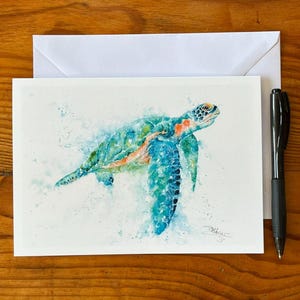 Puede incluir: Una pintura de acuarela de una tortuga marina en tonos azules, verdes y naranjas, presentada en una tarjeta blanca con un sobre a juego y un bolígrafo negro. La obra de arte presenta una firma en la esquina inferior derecha de la tarjeta.