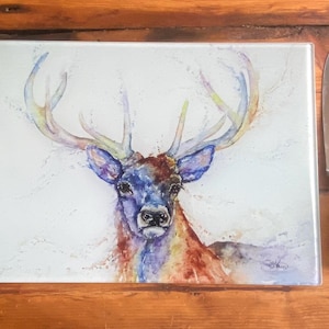 Puede incluir: Tabla de cortar rectangular de vidrio con una pintura de acuarela de un ciervo. El ciervo tiene pelaje azul, morado y rojo con grandes astas. Un cuchillo de acero inoxidable con mango negro está al lado.