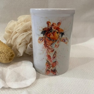 May include: White ceramic tumbler featuring a watercolor giraffe design. The giraffe has orange, brown, and red tones. The tumbler is cylindrical and appears to be a bathroom accessory. Other items in the image include cotton pads and a loofah.