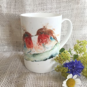 Puede incluir: Taza de cerámica blanca con una ilustración de acuarela de dos petirrojos posados en una rama. Los pájaros se miran y parecen estar cantando.