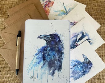 May include: A set of watercolor greeting cards featuring various animals, including a raven, a dragonfly, a badger, and a rabbit. The cards are packaged in a white metal tin with a blue and white watercolor raven design.
