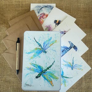 May include: A set of six blank greeting cards featuring watercolour illustrations of dragonflies, a bumblebee, a thistle, and a rabbit. The cards are packaged in a brown envelope with a black pen.