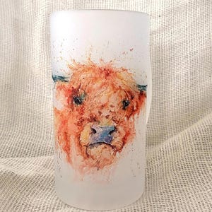 Puede incluir: Un vaso de vidrio esmerilado con una pintura de acuarela de una vaca de las Tierras Altas. La vaca tiene pelaje naranja y marrón, una nariz azul y ojos oscuros. El vaso es cilíndrico.