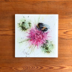 Puede incluir: Pieza de arte de vidrio cuadrada con una pintura de acuarela de un abejorro sobre una flor de cardo rosa. La obra de arte tiene acentos verdes y blancos, con una firma en la esquina inferior izquierda. Colocada sobre un fondo de madera.