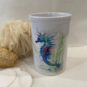 Puede incluir: Vaso de cerámica blanca con un diseño de caballito de mar colorido. El caballito de mar está representado en tonos azules, morados y verdes, con un efecto acuarela. El vaso es cilíndrico y parece un accesorio de baño.