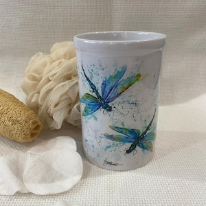 May include: White ceramic tumbler with a watercolour design of blue and teal dragonflies. The tumbler is cylindrical and features a speckled background. Other items in the image include cotton pads and a loofah.