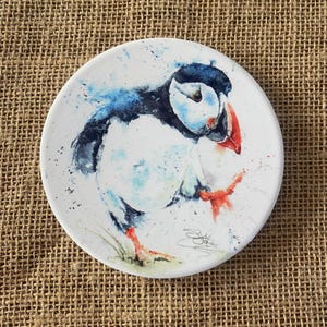 May include: A round, white ceramic coaster featuring a watercolor painting of a puffin. The bird has a black head, white body, and a bright red and orange beak and feet. Blue watercolor splashes surround the puffin.
