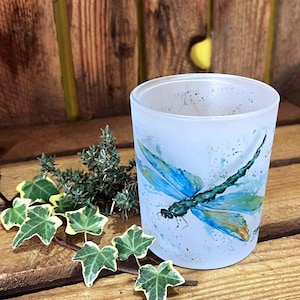 May include: A frosted glass candle holder featuring a watercolour-style illustration of a dragonfly with blue and green wings. The design includes paint splatters and a small signature. A sprig of green ivy with variegated leaves rests nearby.