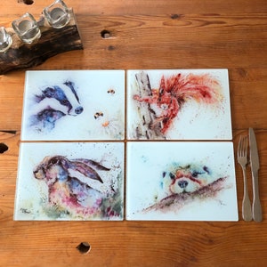 May include: Set of four square glass coasters with watercolour illustrations of a badger, squirrel, hare, and otter. Each coaster features a white background with a light blue, pink, and green watercolour design.