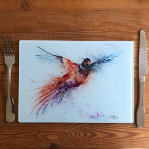 Peut inclure: Planche à découper rectangulaire en verre avec une peinture à l'aquarelle d'un oiseau en vol. L'oiseau présente des nuances de rouge, d'orange, de violet et de bleu. Une fourchette et un couteau sont de chaque côté de la planche, posés sur une surface en bois.