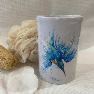 Könnte beinhalten: Weißer Keramikbecher mit einem lebendigen Aquarellgemälde eines blauen und gelben Vogels im Flug. Der Becher ist von einem Luffa, Wattepads und einem beigefarbenen Waschlappen umgeben, was auf ein Badezimmer- oder Spa-Thema hindeutet.