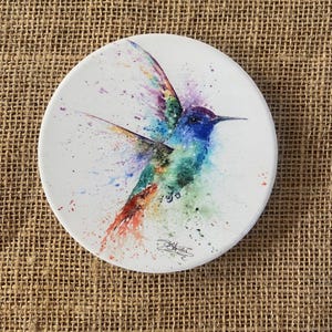 May include: A white ceramic coaster with a watercolor painting of a hummingbird in flight. The hummingbird is painted in shades of blue, green, purple, and orange. The background is white with splashes of color.