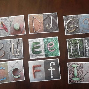 May include: Nine cards with different letters of the alphabet made from natural materials. Each card has a different background and the letters are made from items like leaves, twigs, and stones.