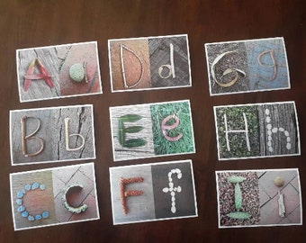 May include: Nine cards with different letters of the alphabet made from natural materials. Each card has a different background and the letters are made from items like leaves, twigs, and stones.