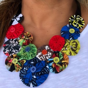 May include: A colorful fabric necklace with a variety of patterned fabric flowers and buttons. The necklace is made of a black cord and is worn by a person.