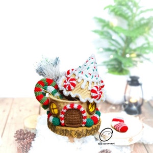 May include: A crocheted gingerbread house with a candy cane and a white and green crocheted tree inside. The house is decorated with red and white candy canes and green sprinkles. The house is sitting on a wooden base.