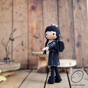 May include: Crocheted doll with black hair in two braids, wearing a black dress with white stripes, black shoes, and a small backpack. The doll has a pale complexion and is standing on a wooden surface.