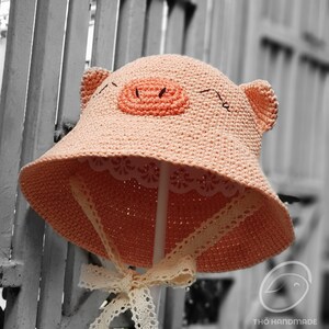 May include: A pink crocheted sun hat shaped like a pig with a white lace trim and a matching white ribbon tied in a bow. The hat is on a white stand against a gray background.