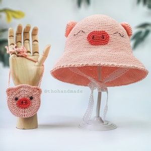 Peut inclure: Un chapeau de cochon rose en crochet avec une bordure en dentelle blanche et un sac &agrave; main en crochet de cochon rose assorti avec une sangle. Le chapeau et le sac &agrave; main sont expos&eacute;s sur un support blanc. Le texte '@thohandmade' est visible sur l'image.
