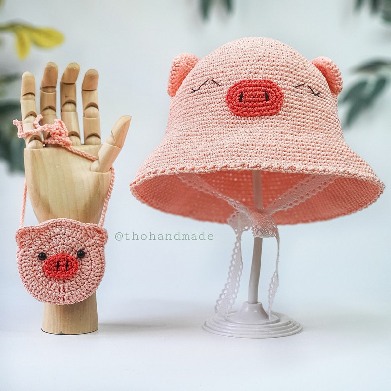 Peut inclure: Un chapeau de cochon rose en crochet avec une bordure en dentelle blanche et un sac &agrave; main en crochet de cochon rose assorti avec une sangle. Le chapeau et le sac &agrave; main sont expos&eacute;s sur un support blanc. Le texte '@thohandmade' est visible sur l'image.