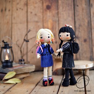 May include: Two handmade crochet dolls, one with blonde hair and a blue dress, the other with black hair and a black dress. Both dolls have detailed facial features and are standing on a wooden surface. The dolls are wearing school uniforms and backpacks.