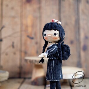 May include: A handmade crochet doll with black hair in braids, wearing a black dress with white stripes and a small backpack. The doll has a fair complexion and is holding a small object. The background is a blurred wooden surface.