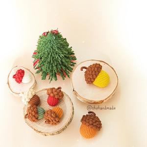 May include: A collection of crocheted acorn ornaments in various colors, including brown, red, yellow, and green. The acorns are displayed on small wooden rounds, with a crocheted Christmas tree in the background. The text '@thohandmade' is visible in the image.