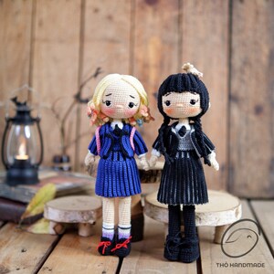 May include: Two handmade crochet dolls, one with blonde hair and the other with black hair, dressed in school uniforms. The blonde doll wears a blue dress with a pink backpack, while the black-haired doll is in a black outfit. Both dolls are holding hands.