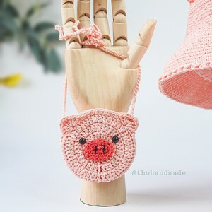 Peut inclure: Un petit sac &agrave; main en crochet rose en forme de cochon avec des yeux noirs et un nez rouge. Le sac est suspendu &agrave; une main en bois.