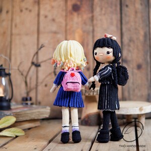 May include: Two handmade crochet dolls, one with blonde hair and a pink backpack, the other with black hair and a black outfit. The dolls are standing on a wooden surface, with a rustic wooden background. The doll's outfits are detailed with blue and black colors.