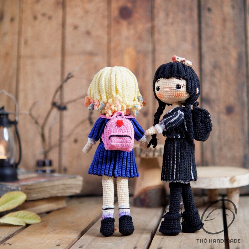 May include: Two handmade crochet dolls, one with blonde hair and a pink backpack, the other with black hair and a black outfit. The dolls are standing on a wooden surface, with a rustic wooden background. The doll's outfits are detailed with blue and black colors.