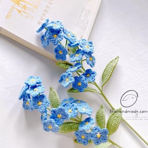 May include: Handmade crochet forget-me-not flowers in blue with yellow centres and green stems and leaves. The flowers are displayed on a white surface, with a book in the background. A delicate craft item.