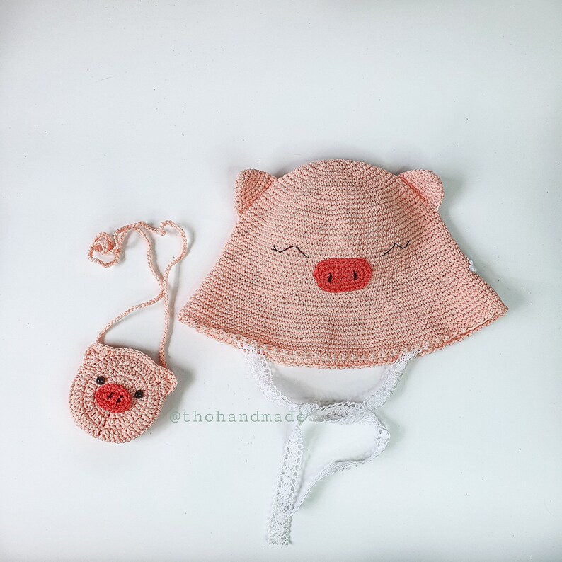 Peut inclure: Un chapeau et un sac &agrave; main assortis en crochet rose avec des visages de cochons. Le chapeau est orn&eacute; d'une bordure en dentelle blanche et de liens. Le sac &agrave; main a une bandouli&egrave;re noire.